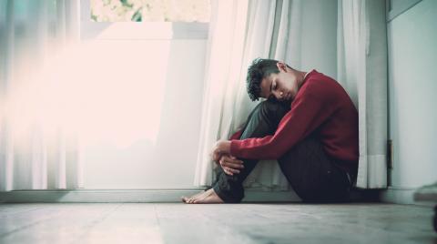 Man sitting hugging his knees near a white wall and window