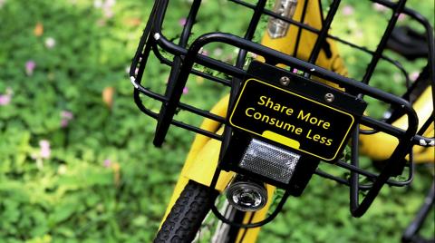 Tackling climate change calls us to a new relationship with nature, and lives of greater simplicity. Photo: Pop Zebra - unsplash.com Yellow bike on green grass with a sign that says 'Share more, consume less'