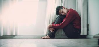 Man sitting hugging his knees near a white wall and window