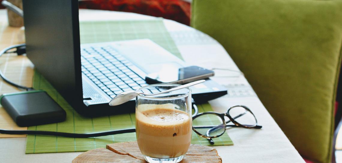 One or two days each week can be regularly worked from home, if your employer agrees. Photo: djurdjica boskovic-unsplash Coffee sitting next to a laptop on a desk with a green chair in the background.