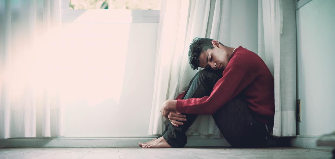 Man sitting hugging his knees near a white wall and window