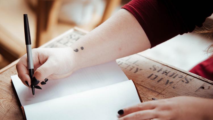 Person writing in a notebook with a semicolon tattoo on their wrist.