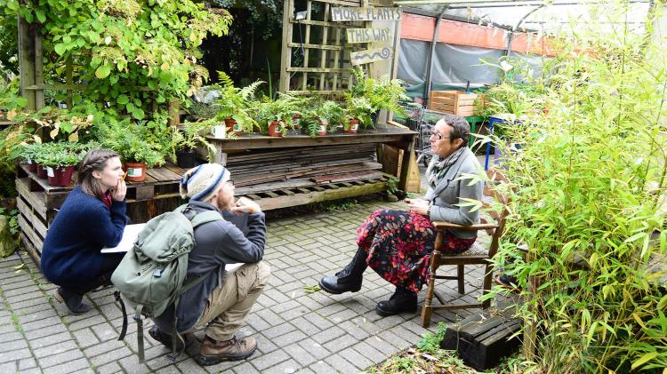 Two people squat while listening to a person sitting in a chair talking. They are all in a garden.