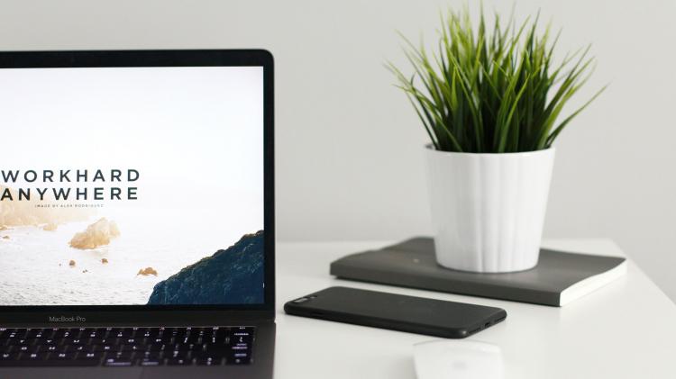 It's impossible to know the exact contribution that trips to meetings and conferences make to carbon emissions. However, it’s estimated that of the 488 million business trips Americans make a year. Photo: Kevin Bhagat - unsplash.com Computer sitting on white desk with phone and house plant. Screen reads "work hard anywhere"