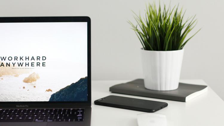 Computer sitting on white desk with phone and house plant. Screen reads "work hard anywhere"